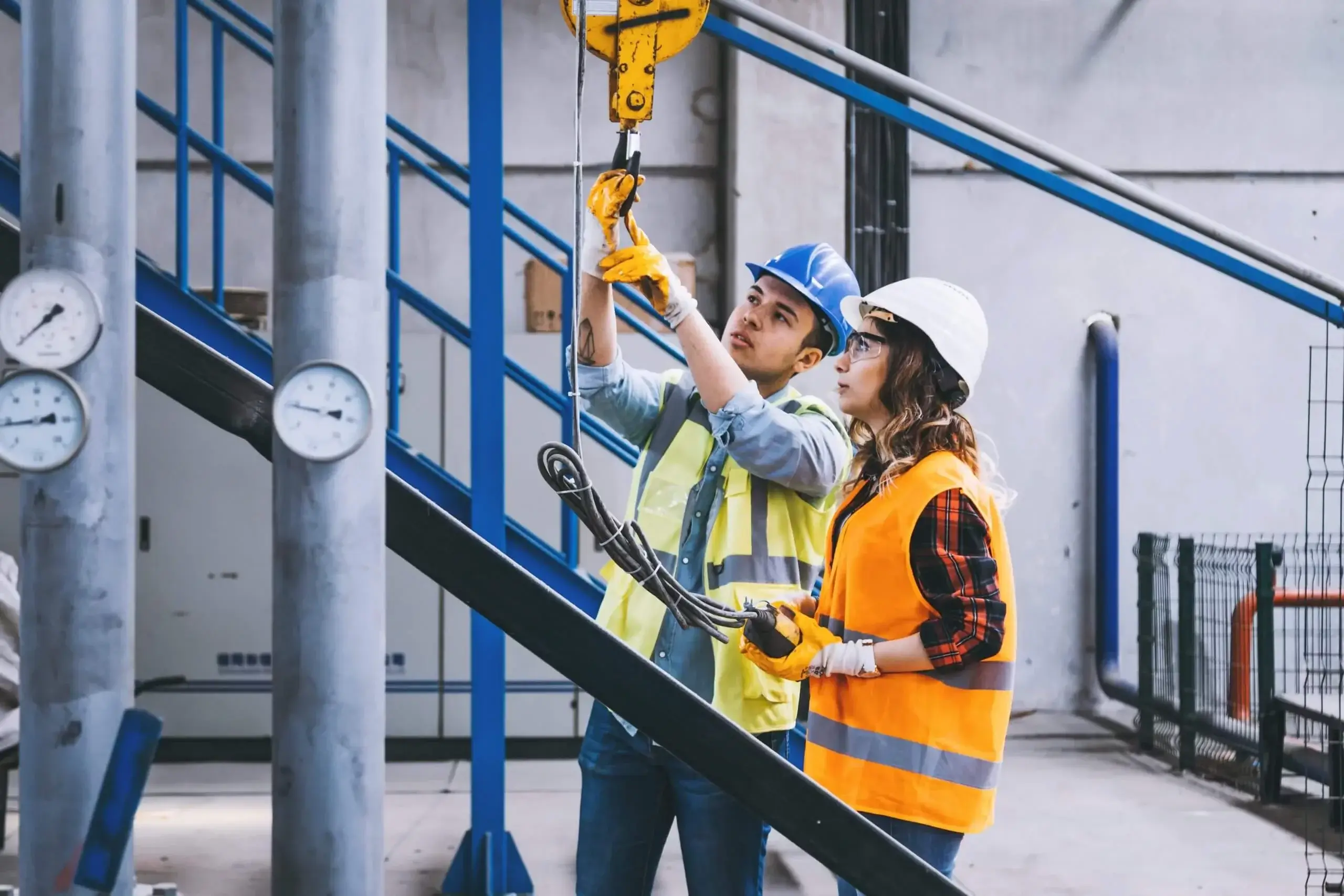 Young heavy industry worker businessman and engineer woman holding crane hook button working with safety workwear and moving a massive metal construction object in warehouse of factory. XXXL size